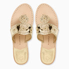 A pair of gold metallic slide sandals with ornate whipstitch rondel medallion details on the strap, shown from a top-down view on a white background. 
