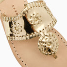 A close-up of a metallic gold thong sandal featuring a wide, whipstitched strap with a rondel medallion loral motifs, set against a light beige leather footbed.