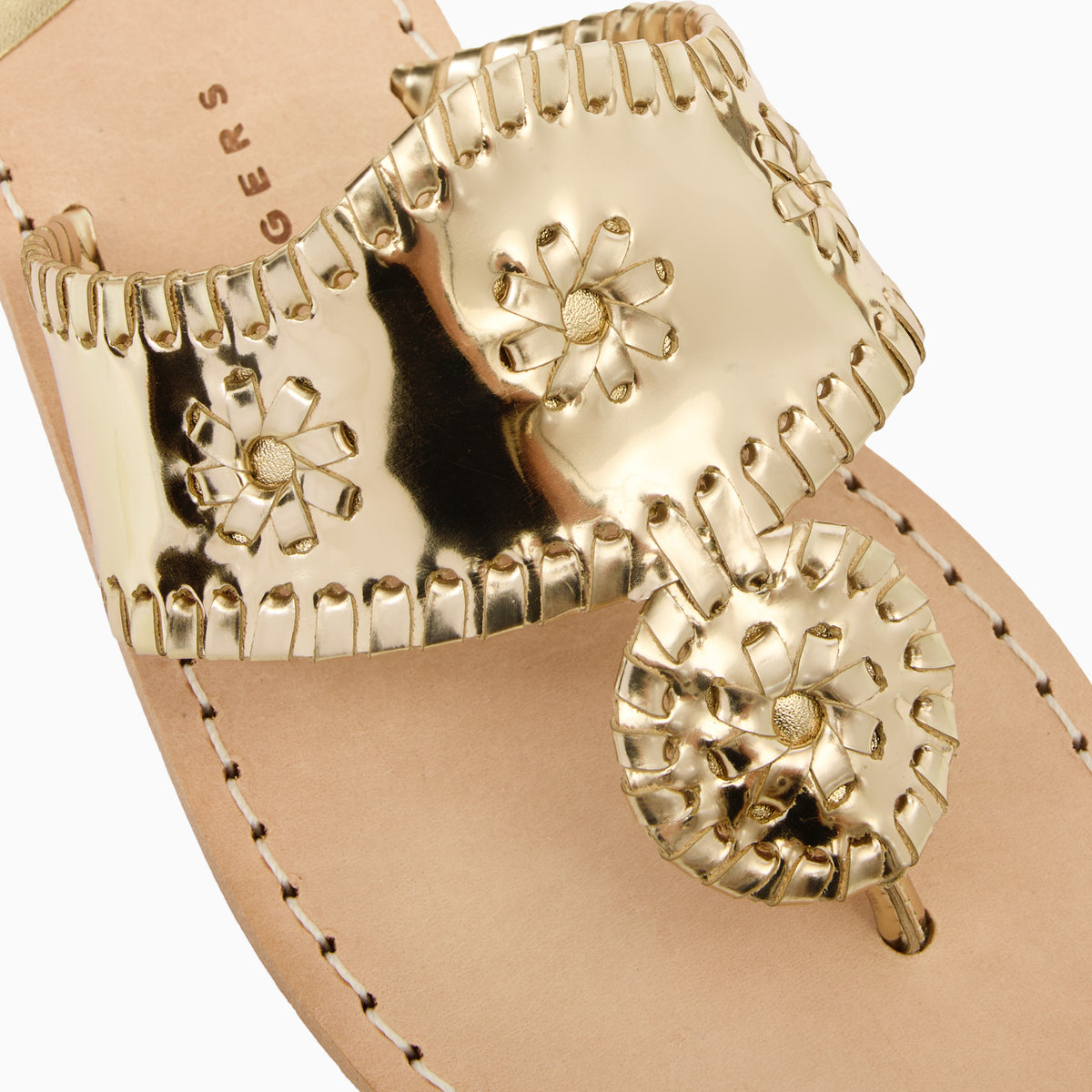 A close-up of a metallic gold thong sandal featuring a wide, whipstitched strap with a rondel medallion loral motifs, set against a light beige leather footbed.